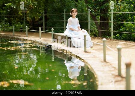 Fille en robe blanche assise sur le rocher près de l'étang dans le jardin Banque D'Images