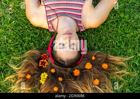 Jeune femme avec les yeux fermés écouter de la musique à travers des écouteurs avec des fleurs dans les cheveux couchés sur l'herbe Banque D'Images