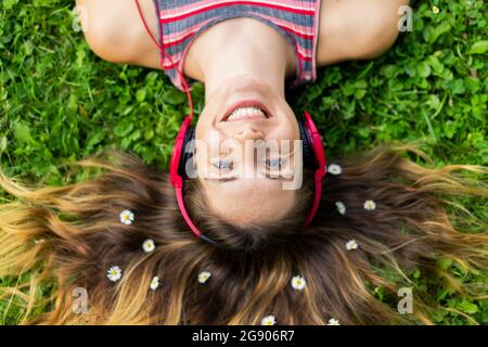 Bonne jeune femme écoutant de la musique avec des écouteurs avec des fleurs dans les cheveux couchés sur l'herbe Banque D'Images