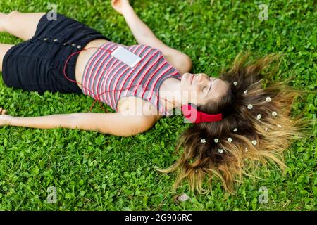 Jeune femme écoutant de la musique à travers des écouteurs avec des fleurs dans les cheveux couchés sur l'herbe Banque D'Images