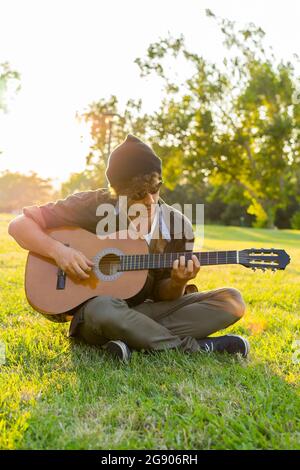 Jeune homme portant un bonnet assis à pattes croisées jouant de la guitare au parc Banque D'Images