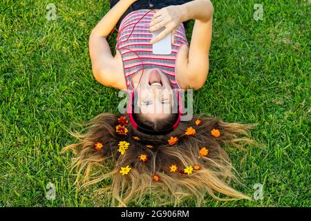 Jeune femme espiègle avec des fleurs dans les cheveux qui collent la langue sur l'herbe Banque D'Images