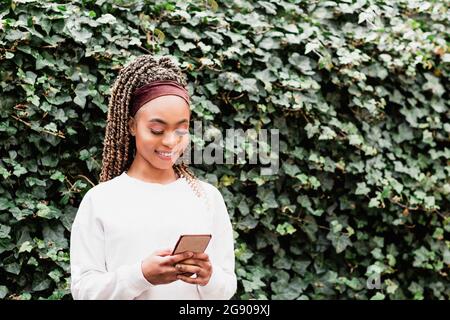 Femme souriante messagerie texte à travers le smartphone debout devant des plantes de lierre vertes Banque D'Images