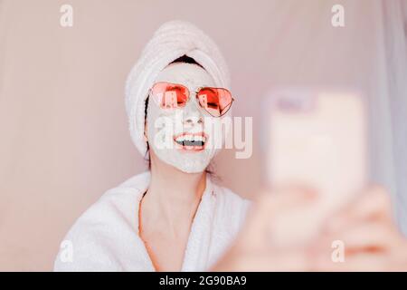 Bonne jeune femme avec masque de beauté prenant le selfie à travers le téléphone intelligent Banque D'Images