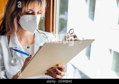 Femme médecin portant un masque facial de protection écrivant sur le presse-papiers en clinique Banque D'Images