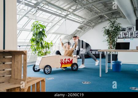 Un collègue qui pousse un homme d'affaires enjoué assis dans une voiture-jouet dans un bureau créatif Banque D'Images