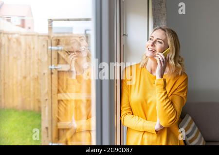 Femme souriante parlant sur un smartphone près de la porte de la maison Banque D'Images