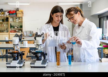 Une scientifique féminine lit le document pendant que la collègue verse du liquide dans un cylindre gradué Banque D'Images