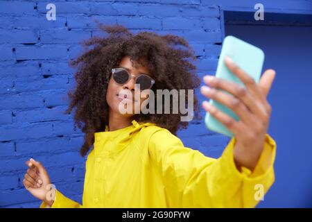 Jeune femme portant des lunettes de soleil emportant un selfie à travers un smartphone devant un mur bleu Banque D'Images