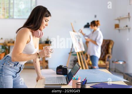 Une photographe féminine tient une tasse penchée sur son bureau tout en peignant un artiste en arrière-plan Banque D'Images