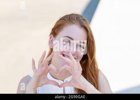 Femme d'affaires souriante avec tête rouge montrant le geste de forme de coeur pendant la journée ensoleillée Banque D'Images