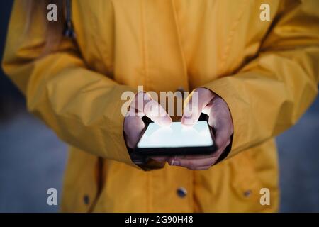 Jeune femme portant un imperméable jaune avec messagerie texte sur un smartphone Banque D'Images