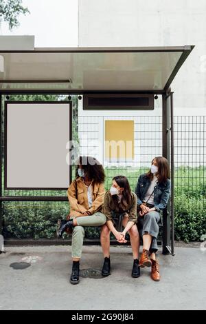 Des amis avec masque facial attendent pendant qu'ils sont assis à l'arrêt de bus Banque D'Images