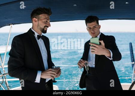 Homme souriant regardant un ami qui prend un selfie en yacht pendant les vacances Banque D'Images