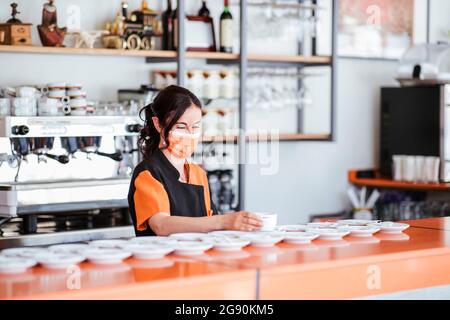 Serveuse portant un masque facial de protection pour organiser une tasse sur la soucoupe au comptoir de la cafétéria Banque D'Images