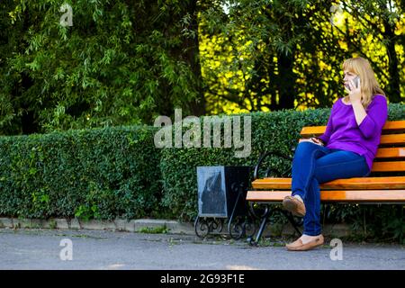 Flou artistique Femme blonde caucasienne parlant, parlant au téléphone à l'extérieur, à l'extérieur. femme de 40 ans en chemisier violet dans le parc sur banc. Femme adulte Banque D'Images