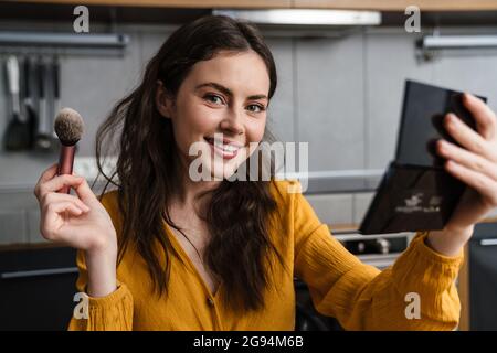 Souriante jeune femme brune faisant le maquillage tout en s'asseyant dans la cuisine à la maison, tenant le miroir et la mascara Banque D'Images