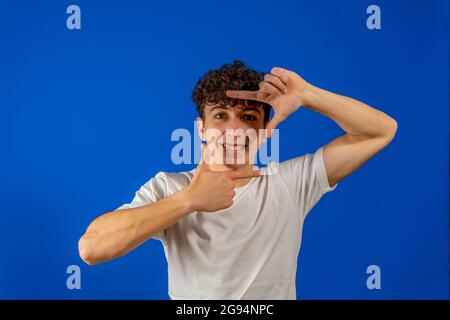 Photo de charmant jeune gars habillé blanc chemise montrant l'appareil photo prise gestuelle sourire isolé couleur bleu arrière-plan. Concept de geste Banque D'Images