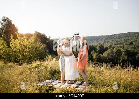 Vue arrière de la compagnie de trois jeunes filles amis s'amuser, boire du vin rouge, élever des verres et profiter du paysage de colline verte et beau coucher de soleil au pique-nique d'été. Banque D'Images