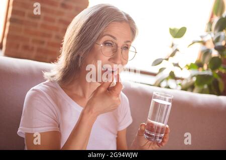 Portrait de la femme attrayante en bonne santé à cheveux gris assis sur divan prenant des pilules médicam à loft maison industrielle à l'intérieur Banque D'Images