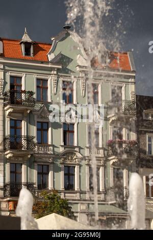 Lumière du soleil du soir sur les façades - vieille ville de Gniezno, Pologne. Fontaine sursaut, gouttes d'eau, rue principale dans la ville comme un fond. Banque D'Images