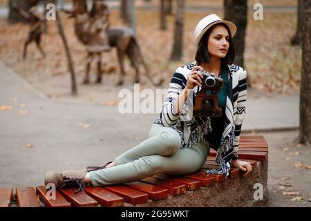 Almaty, Qazaqstan - 02 mai 2021: Jeune belle femme dans un chapeau avec une caméra rétro se trouve sur un banc dans un parc d'automne Banque D'Images