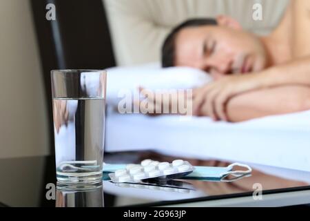 Homme malade dormant dans un lit, des pilules et du verre d'eau au premier plan. Concept de maladie, fièvre, symptômes du coronavirus Banque D'Images