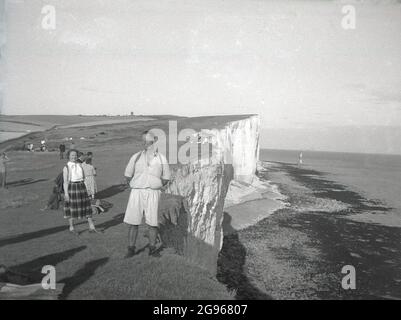 Années 1950, historique, de jour et un marcheur mâle en short debout pour une photo près - très près du bord de la falaise à Beachy Head, un bout de craie sur les South Downs, à Eastbourne, East Sussex, Angleterre, Royaume-Uni. La plus haute falaise de la mer de craie en Grande-Bretagne, les vallons environnants sont un endroit célèbre pour la marche de plaisir. Une zone d'une beauté naturelle exceptionnelle, en 1929, une loi du Parlement a permis d'acheter les terres afin de protéger leur utilisation pour les générations futures. Le phare offshore construit en 1902 peut être vu au loin. Banque D'Images