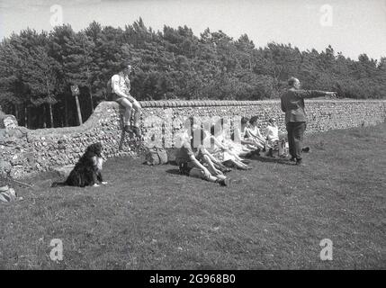 1950s, historique, un groupe de Ramblers se reposant dans un champ à côté d'un mur de pierre à côté d'une forêt sur le sud Downs, East Sussex, Angleterre, Royaume-Uni, obtenant quelques directions d'un gentleman dans un costume avec son pantalon inséré dans ses chaussures. Banque D'Images