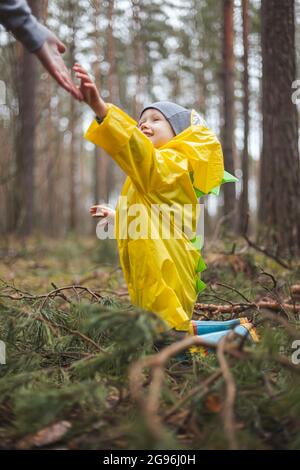 Enfant dans un imperméable jaune marchant dans la forêt après la pluie, assis sur le sol dans les branches de pin, maman atteint et l'aide à se lever Banque D'Images