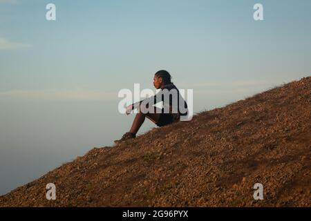 Uribia, la Guajira, Colombie - Mai 28 2021: Jeune homme brun latin avec des braïdes sur sa tête est assis pensivement en attendant le coucher du soleil dans le désert Banque D'Images