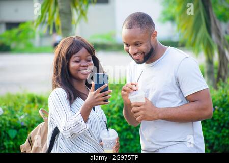 Des amis africains composés d'un gars et d'une dame qui interagissent joyeusement ensemble en regard du smartphone dans la main de la dame Banque D'Images