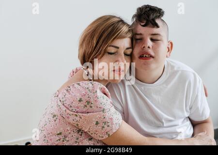 Mère embrassant un garçon handicapé de 17 ans en fauteuil roulant avec paralysie cérébrale sur fond blanc. Femme aide la personne ayant des besoins spéciaux. Émotionnel Banque D'Images