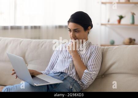 Femme indienne attentionnés regardant l'écran d'un ordinateur portable, assise sur un canapé Banque D'Images