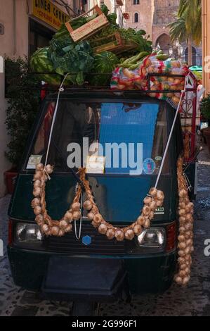 Des cordes ou des cordes d'oignons sont enroulés sur l'unique essuie-glace central de ce piaggio APE pick-up à trois roues, empilé haut avec des boîtes de fruits et légumes comme il livre aux détaillants en ombré via Mandrilisca sous la façade de la cathédrale normande à Cefalu, Sicile, Italie. Les tricycles motorisés APE (Bee) fabriqués par Piaggio ont un avantage majeur par rapport aux fourgons de livraison conventionnels dans les villes historiques italiennes : un cercle de braquage beaucoup plus serré leur permettant de négocier des ruelles et ruelles médiévales étroites. Banque D'Images