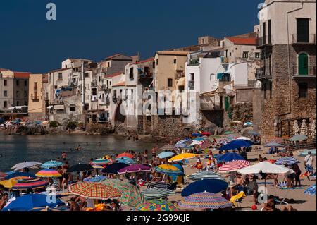 Des corps bronzés et des parasols de plage éclatants - des bandes sedate mêlant fleurs, palmiers et une touche de tartan - remplissent un coin de la plage de la vieille ville à Cefalu, Sicile, Italie, sur fond de murs blancs et ocre de hauts, maisons carrelées de rouge construites sur des vestiges voûtés du mur médiéval de la ville. La ville de la côte nord, fondée par les Grecs anciens et occupée à son tour par les Romains, les Byzantins, les Arabes et les Normands, est l'une des stations les plus populaires de Sicile. Banque D'Images