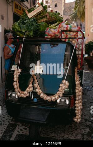 Cordes ou ficelles d'oignon enroulés sur le unique essuie-glace central d'un piaggio APE pick-up à trois roues empilé haut avec des boîtes de fruits et de légumes comme il livre aux détaillants en ombré via Mandrilisca sous la façade de la cathédrale normande à Cefalu, Sicile, Italie. Les tricycles motorisés APE (Bee) fabriqués par Piaggio ont un avantage majeur par rapport aux fourgons de livraison conventionnels dans les villes historiques italiennes : un cercle de braquage beaucoup plus serré leur permettant de négocier des ruelles et ruelles médiévales étroites. Banque D'Images