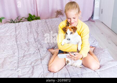 Une adolescente avec un téléphone mobile, embrassant un chien Jack Russell Terrier, communique sur les réseaux sociaux ou via Zoom avec des amis ou la famille, fait un Banque D'Images