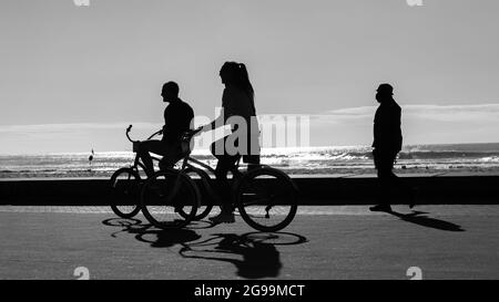 Le couple de vélo silhouetted homme femme non reconnaissable à vélo sur la promenade du front de mer avec le fond des vagues de l'océan en noir et blanc photo. Banque D'Images