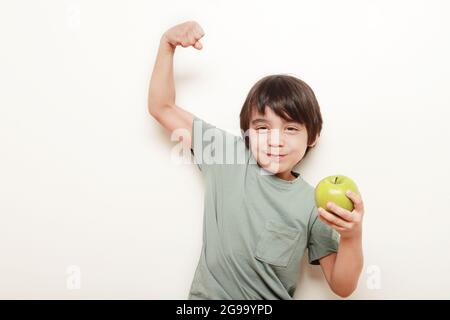 enfant sur fond blanc mangeant la pomme verte qu'il tient d'une main et de l'autre, il nous montre son biceps indiquant la force Banque D'Images