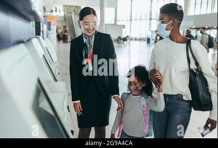 Un employé de compagnies aériennes aide une fille et une mère à utiliser un appareil d'enregistrement en libre-service. Une famille africaine portant un masque facial à l'aéroport avec le personnel des compagnies aériennes. Banque D'Images