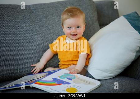 Un adorable enfant australien jouant avec un livre coloré sur un canapé dans une maison Banque D'Images