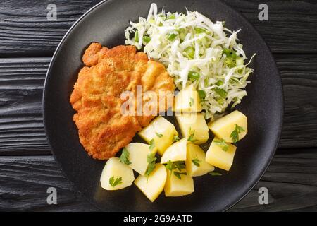 Côtelettes de porc panées polonaises Kotlet Schabowy avec pommes de terre bouillies et salade de chou dans une assiette sur une table en bois noir. Vue de dessus horizontale fr Banque D'Images