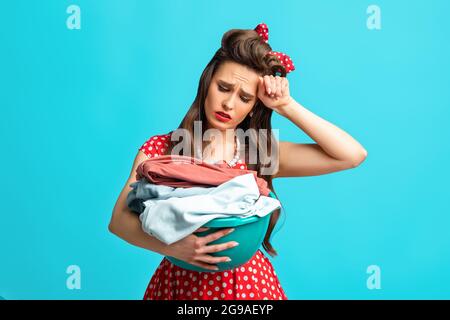 Triste femme épuisée pinup dans une tenue rétro tenant des vêtements pour laver ou repasser sur fond bleu studio Banque D'Images