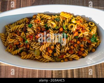 Gros plan de plat de pâtes : fusilli aux pois chiches, câpres, olives et tomates servis dans un plat de cocotte blanc sur une table extérieure au soleil avec un fond de vin Banque D'Images