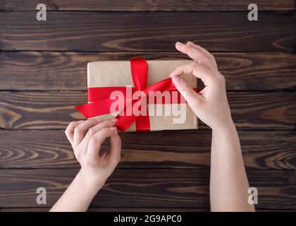 Création d'un noeud dans une boîte cadeau. Femme emballant des vacances fait main présent en papier avec ruban rouge sur table en bois, vue de dessus Banque D'Images
