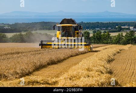 Byres Farm, East Lothian, Écosse, Royaume-Uni, 25 juillet 2021. Météo au Royaume-Uni: Récolte d'orge d'hiver: Le récent temps sec a permis à un agriculteur de commencer tôt la récolte. Photo : une moissonneuse-batteuse New Holland au travail avec vue sur Firth of Forth et Lomond Hills au loin Banque D'Images