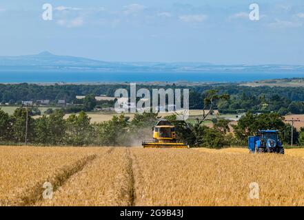 Byres Farm, East Lothian, Écosse, Royaume-Uni, 25 juillet 2021. Météo au Royaume-Uni: Récolte d'orge d'hiver: Le récent temps sec a permis à un agriculteur de commencer tôt la récolte. Photo : une moissonneuse-batteuse New Holland au travail avec vue sur Firth of Forth et Lomond Hills au loin Banque D'Images