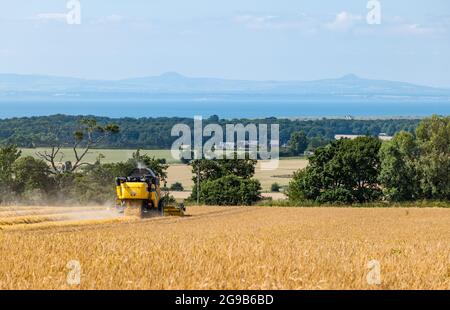 Byres Farm, East Lothian, Écosse, Royaume-Uni, 25 juillet 2021. Météo au Royaume-Uni: Récolte d'orge d'hiver: Le récent temps sec a permis à un agriculteur de commencer tôt la récolte. Photo : la moissonneuse-batteuse est au travail avec une vue sur les collines Firth of Forth et Lomond au loin Banque D'Images