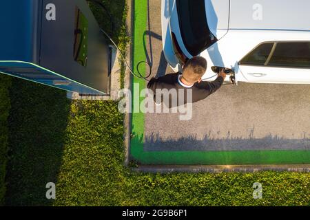 Vue d'oiseau d'un jeune homme du Caucase qui charge une voiture électrique blanche à une station de charge publique Banque D'Images